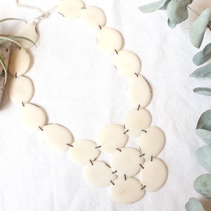 May include: Cream-coloured statement necklace made of oval and circular pieces, joined by dark thread. The necklace is displayed on a white surface, with a sprig of eucalyptus and a small plant in the background.