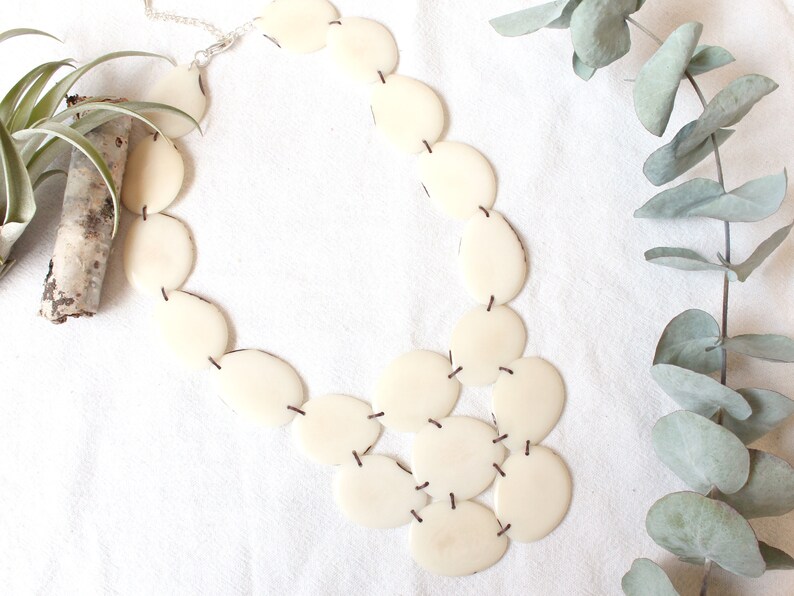 May include: Cream-coloured statement necklace made of oval and circular pieces, joined by dark thread. The necklace is displayed on a white surface, with a sprig of eucalyptus and a small plant in the background.