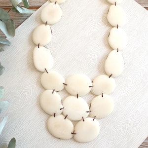 May include: A cream-coloured necklace featuring large, oval-shaped beads strung together with dark brown string. The beads have a smooth, polished appearance. The necklace is displayed on a light-coloured, patterned surface, suggesting a jewelry or accessory theme.