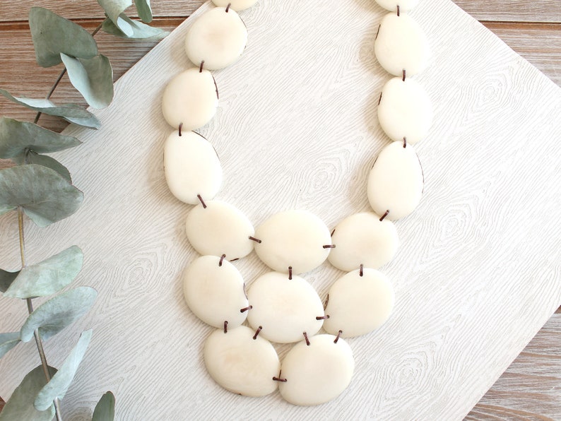 May include: A cream-coloured necklace featuring large, oval-shaped beads strung together with dark brown string. The beads have a smooth, polished appearance. The necklace is displayed on a light-coloured, patterned surface, suggesting a jewelry or accessory theme.