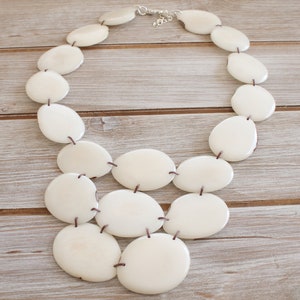 May include: A cream-coloured statement necklace with large, oval-shaped beads. The beads are connected with dark brown string and a silver-toned clasp. The necklace is displayed on a light-coloured wooden surface.