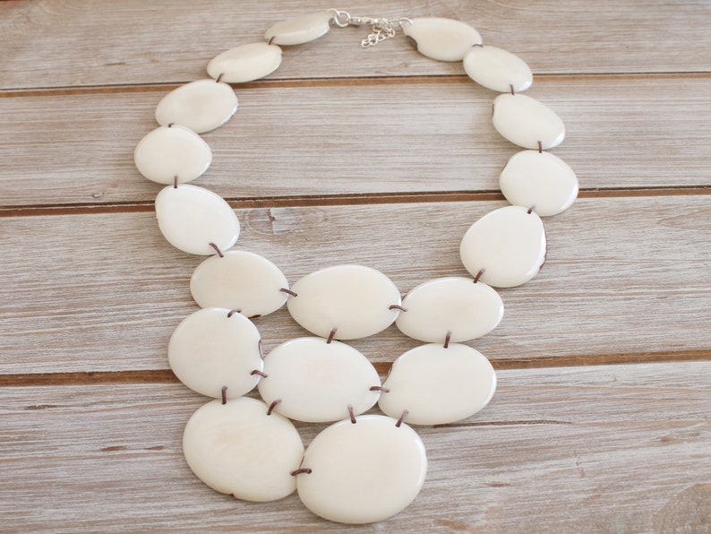 May include: A cream-coloured statement necklace with large, oval-shaped beads. The beads are connected with dark brown string and a silver-toned clasp. The necklace is displayed on a light-coloured wooden surface.