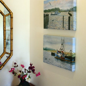 May include: Two framed canvas paintings of coastal scenes hang on a cream-colored wall. The top painting depicts a boat on the water with a bird perched on a wooden post. The bottom painting shows a fishing boat.