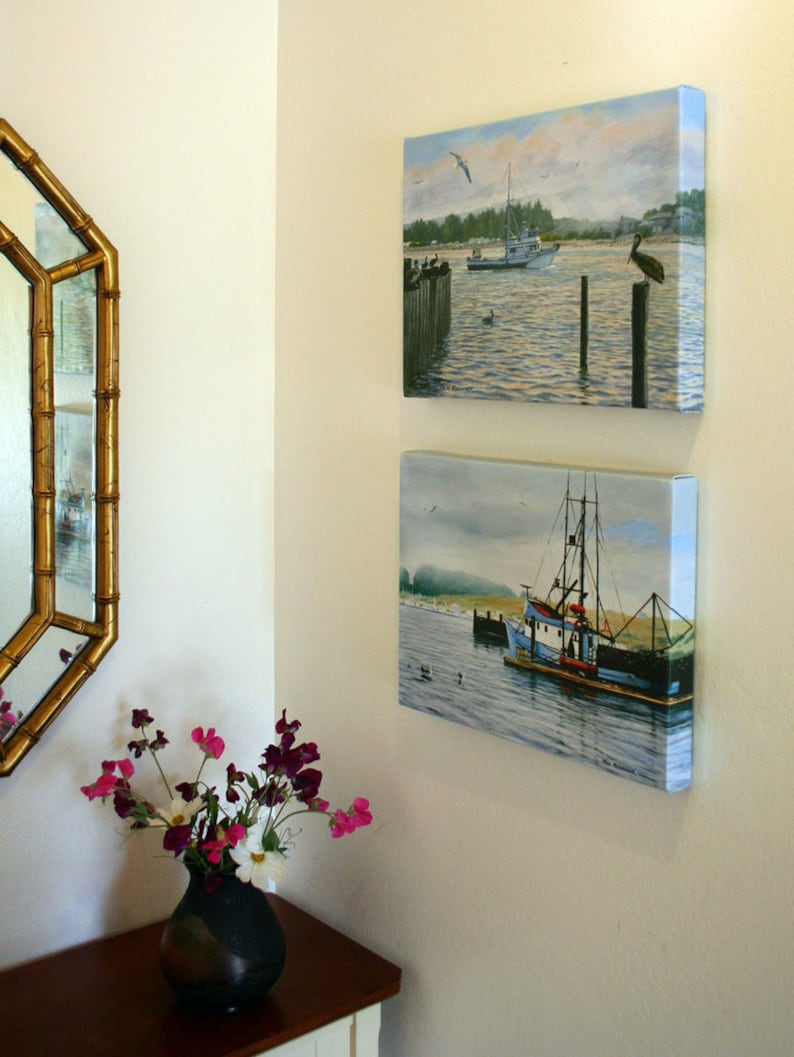 May include: Two framed canvas paintings of coastal scenes hang on a cream-colored wall. The top painting depicts a boat on the water with a bird perched on a wooden post. The bottom painting shows a fishing boat.