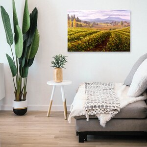 May include: A painting of a vineyard with rows of grapevines in the foreground, a small town in the distance, and a mountain range in the background. The sky is a light blue with white clouds. The painting is hanging on a white wall in a living room setting.