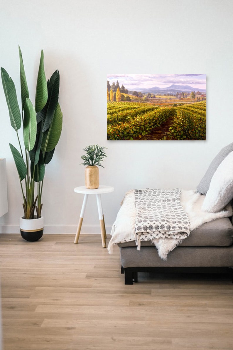 May include: A painting of a vineyard with rows of grapevines in the foreground, a small town in the distance, and a mountain range in the background. The sky is a light blue with white clouds. The painting is hanging on a white wall in a living room setting.