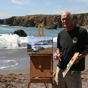 May include: A man paints a seascape on an easel on a beach. The painting depicts a rocky coastline with waves crashing on the shore. The man is wearing a black t-shirt and khaki shorts.