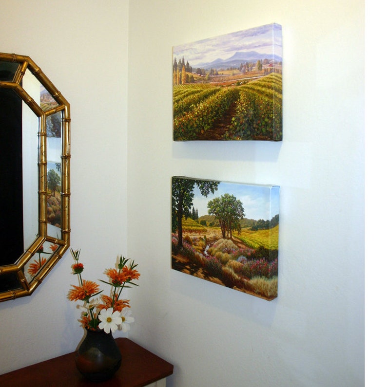 May include: Two framed landscape paintings on a white wall. The top painting shows a vineyard with rolling hills and a distant mountain range. The bottom painting shows a stream running through a meadow with trees and wildflowers.