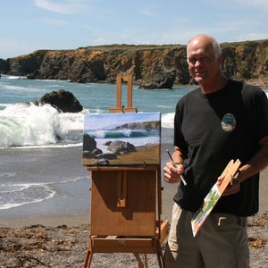 May include: A man paints a seascape on a wooden easel on a beach. The painting depicts a rocky coastline with crashing waves. The man is holding a paintbrush in his right hand and a palette in his left hand.