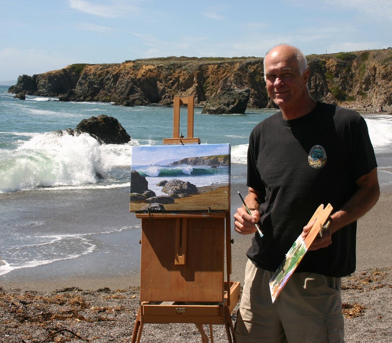 May include: A man paints a seascape on a wooden easel on a beach. The painting depicts a rocky coastline with crashing waves. The man is holding a paintbrush in his right hand and a palette in his left hand.