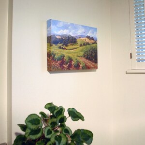 May include: A painting of a rural landscape with rolling hills, green fields, and a blue sky with white clouds. The painting is framed and hanging on a white wall.
