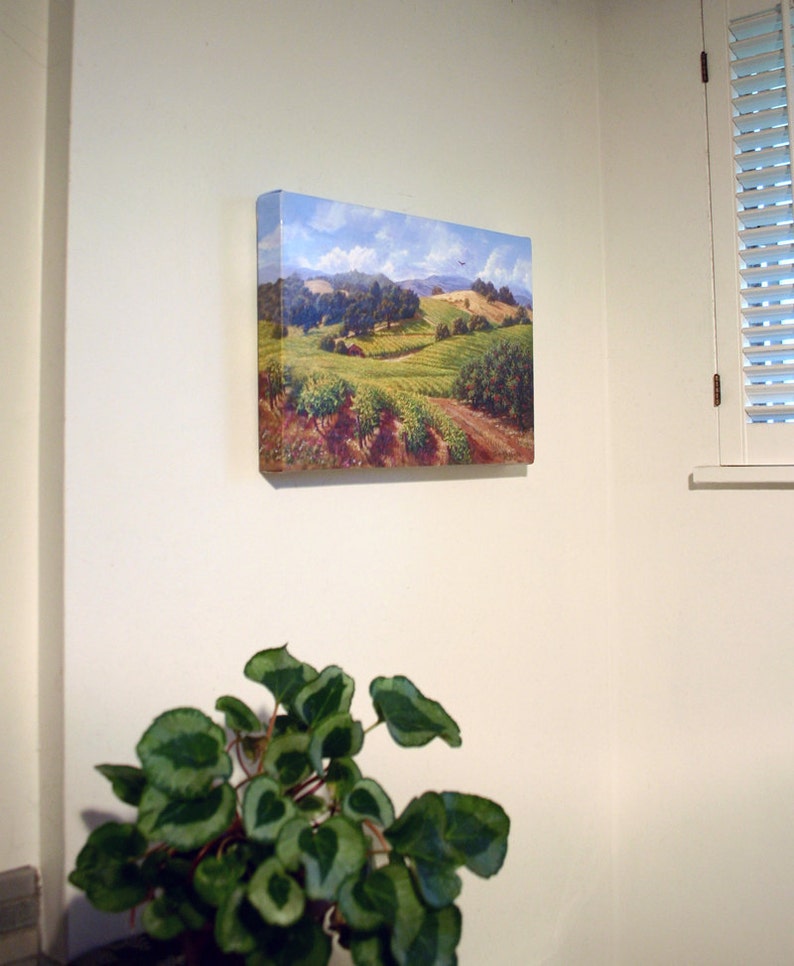 May include: A painting of a rural landscape with rolling hills, green fields, and a blue sky with white clouds. The painting is framed and hanging on a white wall.