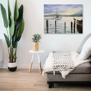 May include: A framed painting depicts a serene coastal scene with a white fishing boat sailing on calm water. Wooden pilings and a dock extend into the water, with birds perched on them. The sky transitions from soft blues to golden hues.