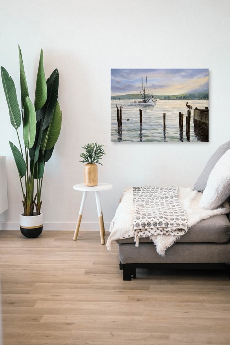 May include: A framed painting depicts a serene coastal scene with a white fishing boat sailing on calm water. Wooden pilings and a dock extend into the water, with birds perched on them. The sky transitions from soft blues to golden hues.