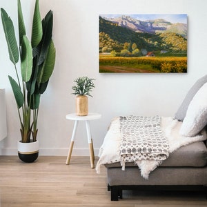 May include: A canvas print showing a vineyard with yellow-green vines, a red-roofed building, and mountains. The artwork hangs on a white wall above a grey sofa with cushions and a throw. A large potted plant and a small side table are in the foreground.