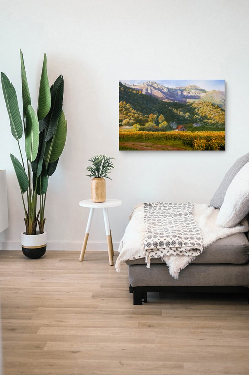May include: A canvas print showing a vineyard with yellow-green vines, a red-roofed building, and mountains. The artwork hangs on a white wall above a grey sofa with cushions and a throw. A large potted plant and a small side table are in the foreground.