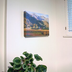 May include: A square canvas art print showing a mountain landscape with a vineyard. The artwork features a red-roofed building, trees, and a mountain range under a blue sky. The print is mounted on a white wall.