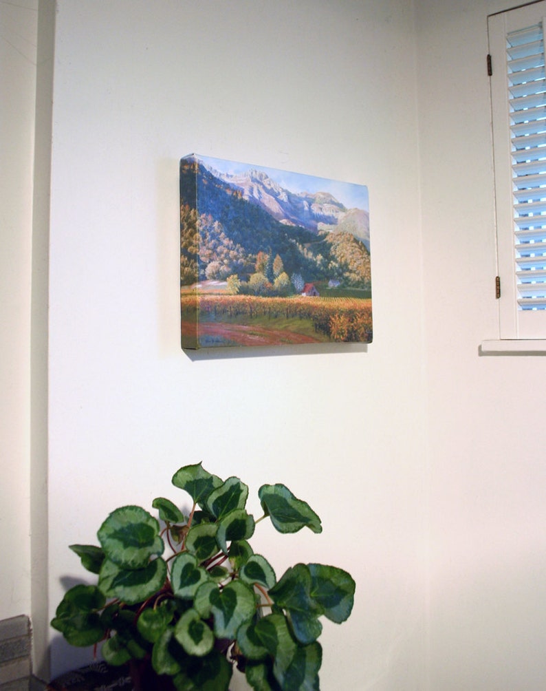 May include: A square canvas art print showing a mountain landscape with a vineyard. The artwork features a red-roofed building, trees, and a mountain range under a blue sky. The print is mounted on a white wall.