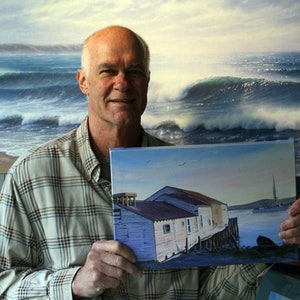 May include: A man holds a painting of a waterfront scene with a group of buildings on stilts. The painting is set against a blue sky with white clouds. The man is wearing a plaid shirt and has a friendly expression.