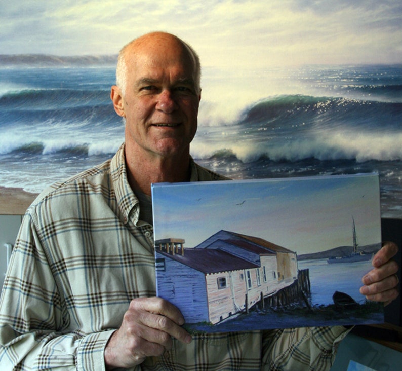 May include: A man holds a painting of a waterfront scene with a group of buildings on stilts. The painting is set against a blue sky with white clouds. The man is wearing a plaid shirt and has a friendly expression.