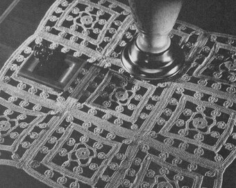 May include: A white crocheted doily with a square design and a floral pattern. The doily is placed on a dark surface with a silver vase in the center.