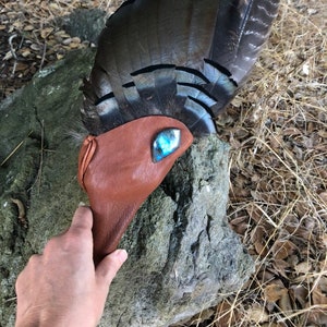 May include: A brown leather pouch with a blue gemstone accent, resting on a rock. The pouch is held by a hand and is next to a large arrangement of dark feathers with black and white patterns. The background features dry grass and foliage.