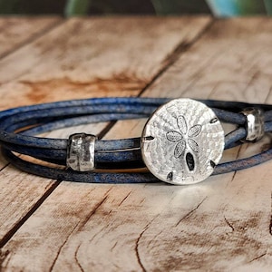 May include: A blue leather wrap bracelet with a silver sand dollar charm. The bracelet is made of multiple strands of leather and has a silver bead accent.