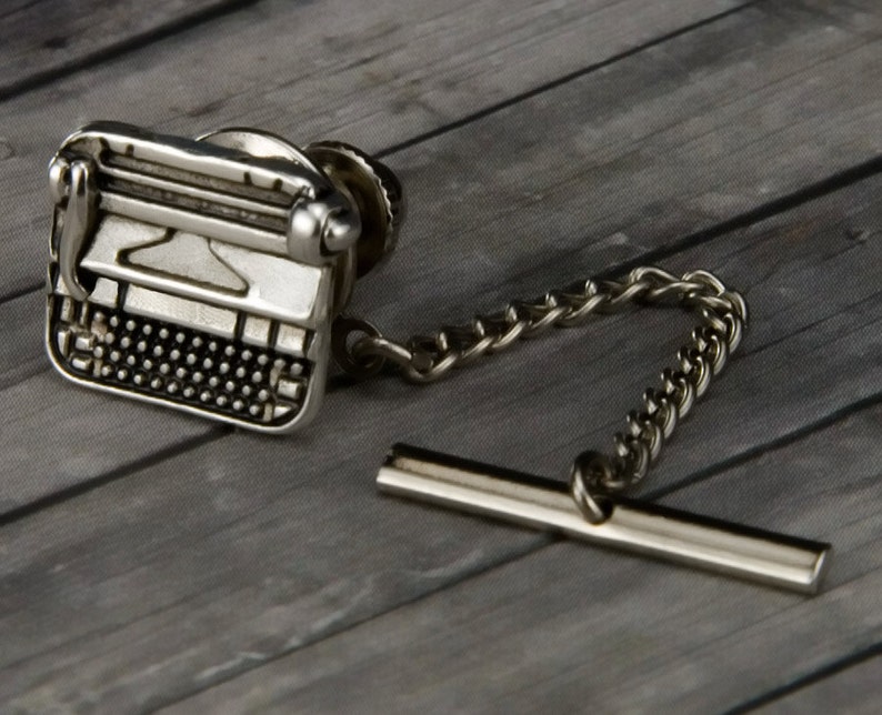 May include: Silver-toned typewriter tie tack with a chain and bar. The tie tack is a miniature replica of a vintage typewriter.