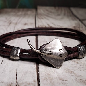 May include: A brown leather bracelet with a silver stingray charm. The bracelet is made of three strands of leather and has two silver beads.