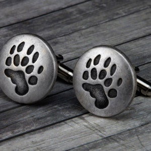 May include: A pair of silver cufflinks with a bear paw print design.