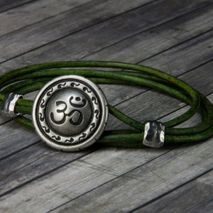 May include: A green leather bracelet with a silver Om symbol charm. The charm is round and has a detailed design.