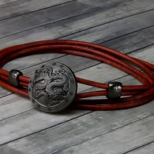 May include: A brown leather bracelet with a black metal dragon charm. The charm is round and has a detailed design of two dragons.