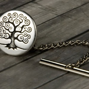 May include: A silver-toned tie tack featuring a detailed tree design within a circular setting. The tree has a textured trunk and swirling branches. A delicate chain and bar complete the accessory, set against a gray wooden background.
