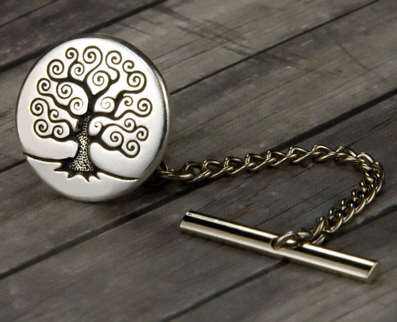 May include: A silver-toned tie tack featuring a detailed tree design within a circular setting. The tree has a textured trunk and swirling branches. A delicate chain and bar complete the accessory, set against a gray wooden background.
