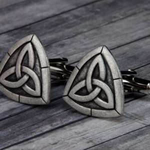 May include: A pair of silver cufflinks with a triangular design featuring a Celtic knot. The cufflinks have a textured finish and a polished silver look.