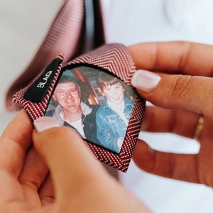 May include: A red and white patterned tie with a small photo of two people inside. The photo shows a man and a younger boy. The tie has a black label with the word "BLAG" on it.