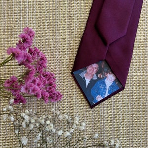 May include: A deep burgundy tie with a concealed photograph of two individuals. The tie rests on a textured, gold-toned surface, accompanied by pink and white dried flowers. The photograph is visible on the reverse side.