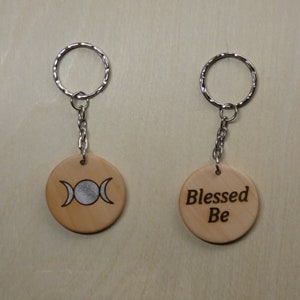 May include: Two wooden keyrings with silver key rings. One keyring features a triple moon symbol and the other says "Blessed Be".