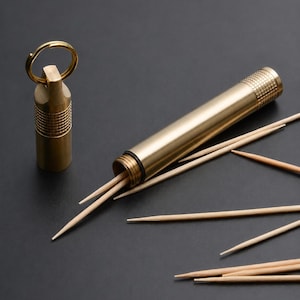 May include: A cylindrical brass toothpick holder with a knurled screw-top cap and an attached metal keyring. The container is open, revealing several light-coloured wooden toothpicks inside, with additional toothpicks scattered around the base.