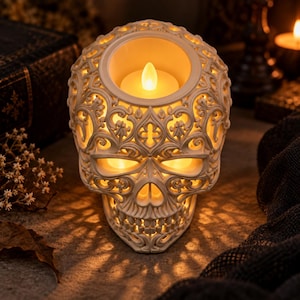 May include: A cream-colored skull-shaped candle holder featuring intricate cut-out filigree patterns across the surface. The top section holds a small, glowing, warm-toned LED tea light, which illuminates the hollow eye sockets and nasal cavity.