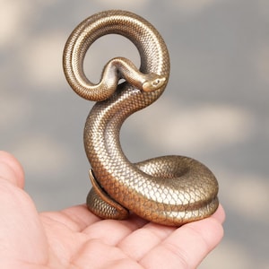 May include: A bronze-coloured snake figurine with a textured, scaled surface. The snake is coiled in an upright, S-shaped position with its head turned slightly to the side. The metallic finish shows subtle highlights and shadows across the scales.