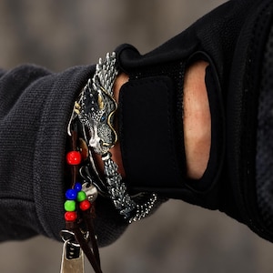 May include: A silver-toned bracelet featuring a detailed dragon head with gold-coloured accents and blue eyes, paired with a brown leather cord bracelet adorned with small red, blue, and green beads and a metallic charm hanging from the end.