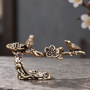 May include: A brass-colored metal figurine depicting two small birds perched on a textured tree branch. The branch features a single open flower blossom and a small cup-like structure on the left side, with a gnarled, root-like base for stability.