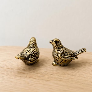 May include: Two small bird figurines crafted from antique-toned brass. Each bird features detailed, etched feather patterns across its body, wings, and tail. One bird is positioned facing away, while the other is turned to the side, showing its beak.
