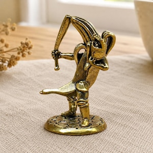 Puede incluir: Una figura de metal color latón que representa un personaje de plátano con casco y armadura, sosteniendo un bate de béisbol sobre el hombro. La figura se apoya sobre una base circular con un patrón texturizado similar a adoquines.