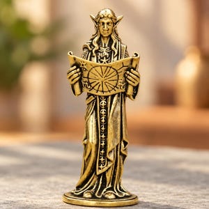 May include: A gold-toned metal figurine of a pointed-eared figure wearing long robes and holding an open scroll. The scroll features a circular star chart design with various symbols. The figure has long hair and stands on a circular base.