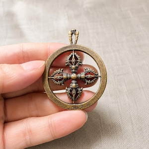 May include: A circular bronze-toned metal pendant featuring a central four-pronged vajra symbol enclosed within a ring frame. The piece has a weathered, antique finish with a small loop at the top for attachment, showcasing intricate detailing.