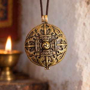 May include: A round, antique gold-toned metal pendant featuring an intricate, embossed vajra or dorje symbol with a central spiral design. The pendant is suspended from a dark brown cord and displays a textured, weathered finish across its surface.