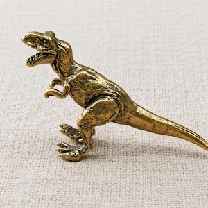 Puede incluir: Una pequeña figura de Tyrannosaurus rex de color oro metálico con un acabado texturizado y desgastado. El dinosaurio se muestra en posición erguida con la boca abierta, mostrando los dientes, brazos pequeños y una larga cola afilada.
