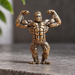Puede incluir: Una pequeña figura metálica de color bronce que representa a un gorila de pie en una pose musculosa con ambos brazos levantados para flexionar los bíceps. La escultura presenta una textura de pelaje detallada, abdominales definidos y una expresión facial estoica.
